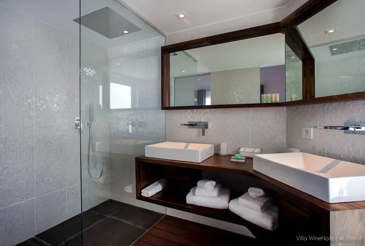 A dark wood double vanity with white vessel sinks sits beneath a large framed mirror, where a walk-in rain shower with frameless glass and warm lighting completes the spa-like space.