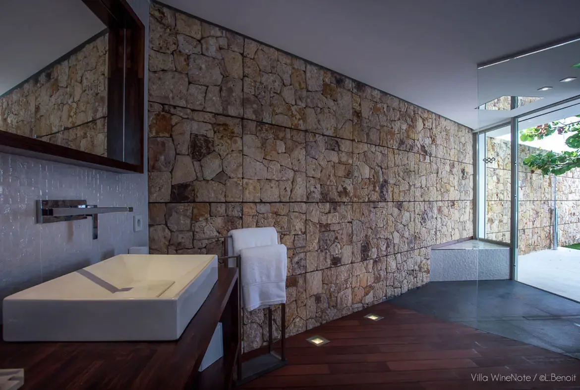 A dark wood vanity with a white vessel sink sits before a natural stone accent wall, where a frameless glass door opens to a sunlit outdoor patio with lush greenery.