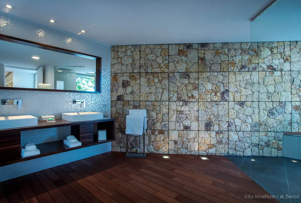 A dark wood floating vanity with two white vessel sinks sits before a textured stone accent wall, where shimmering mosaic tiles and a frameless glass shower create a spa-like retreat.
