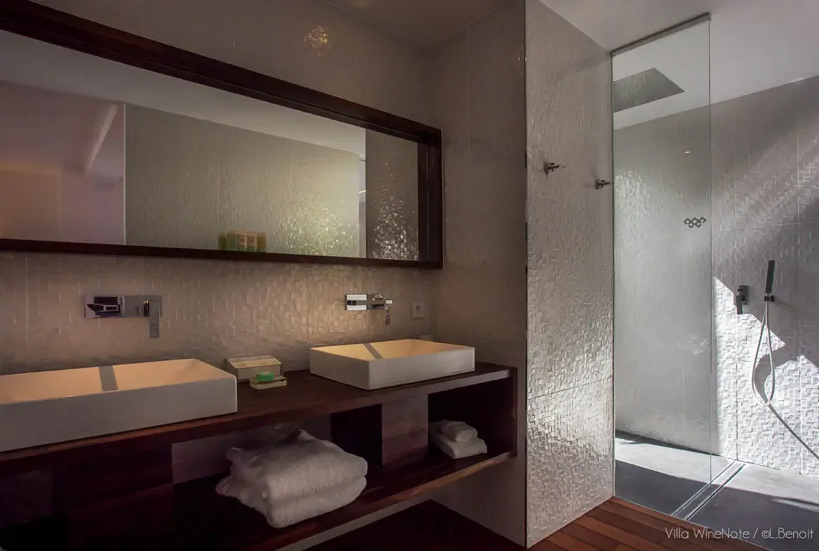A dark wood floating vanity with two white vessel sinks sits beneath a large framed mirror, where shimmering mosaic tiles and a frameless glass shower create a serene spa bathroom.