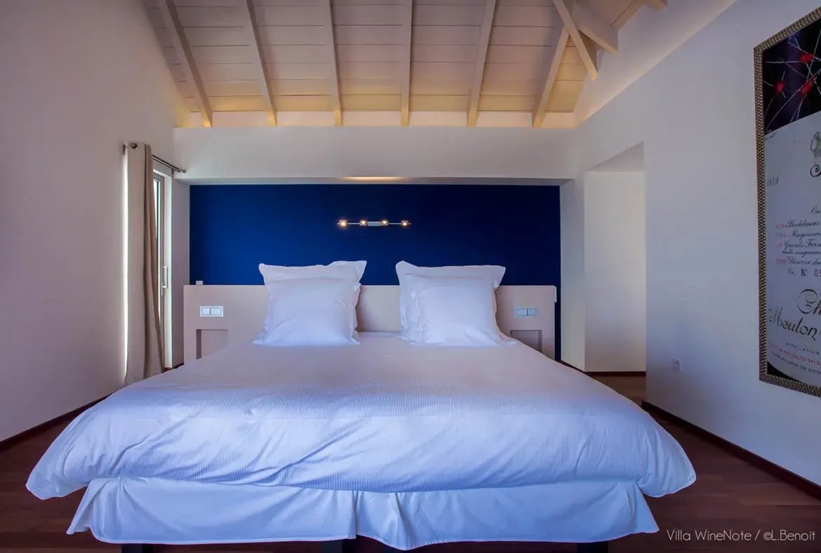 A white bed with plush pillows sits beneath a navy blue accent wall with warm sconces, where a framed wine label artwork hangs above a light-wood headboard in a vaulted-beam bedroom.