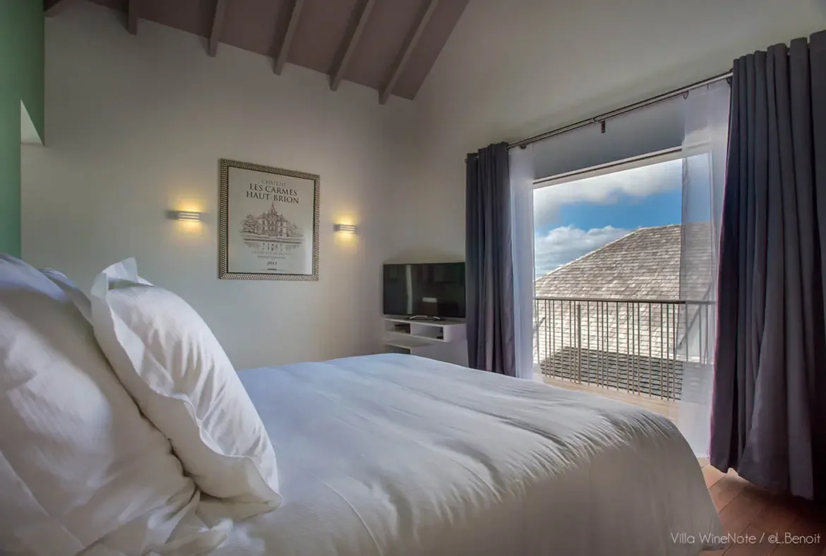 A white bed with plush pillows sits beneath a framed Château Les Carmes Haut-Brion wine label, where warm sconces and vaulted grey-beam ceilings accent the serene bedroom.