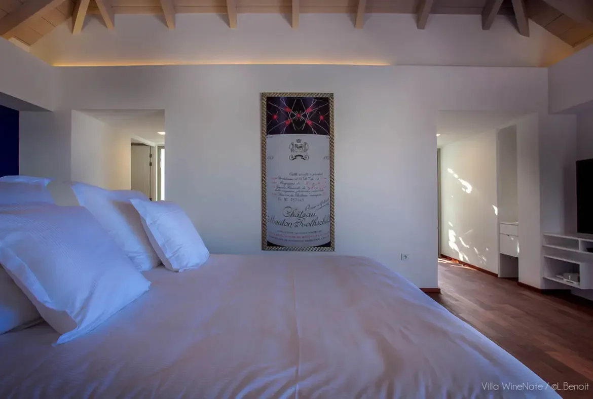 A white bed with plush pillows sits before a framed Château Mouton Rothschild wine label on a white wall, where warm cove lighting and vaulted wood beams accent the serene bedroom.