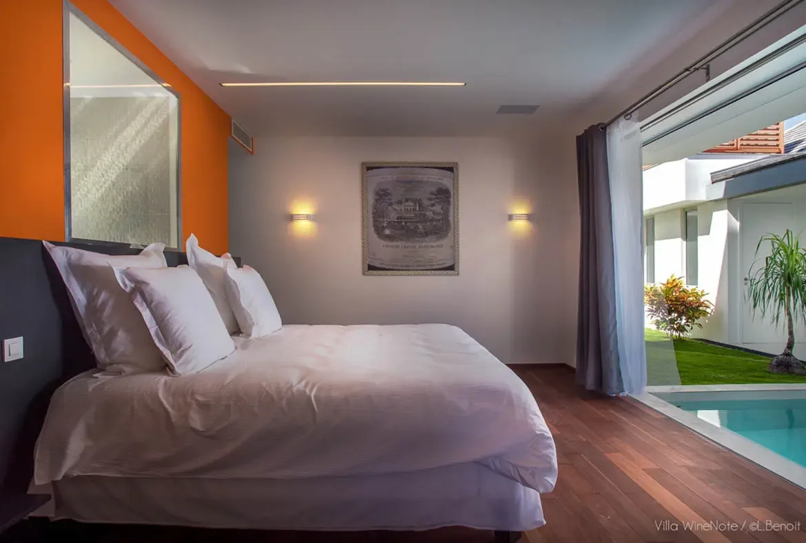A white bed with plush pillows sits before a vibrant orange accent wall and a framed Château Lafite wine label, where open doors lead to a turquoise pool and lush garden.