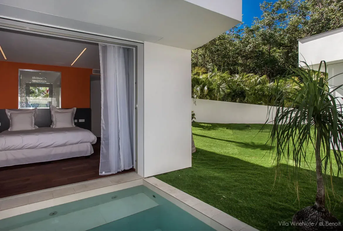 A white bed beside an orange accent wall opens to a turquoise plunge pool with stone stepping stones, where a manicured lawn and tropical garden create a private oasis.