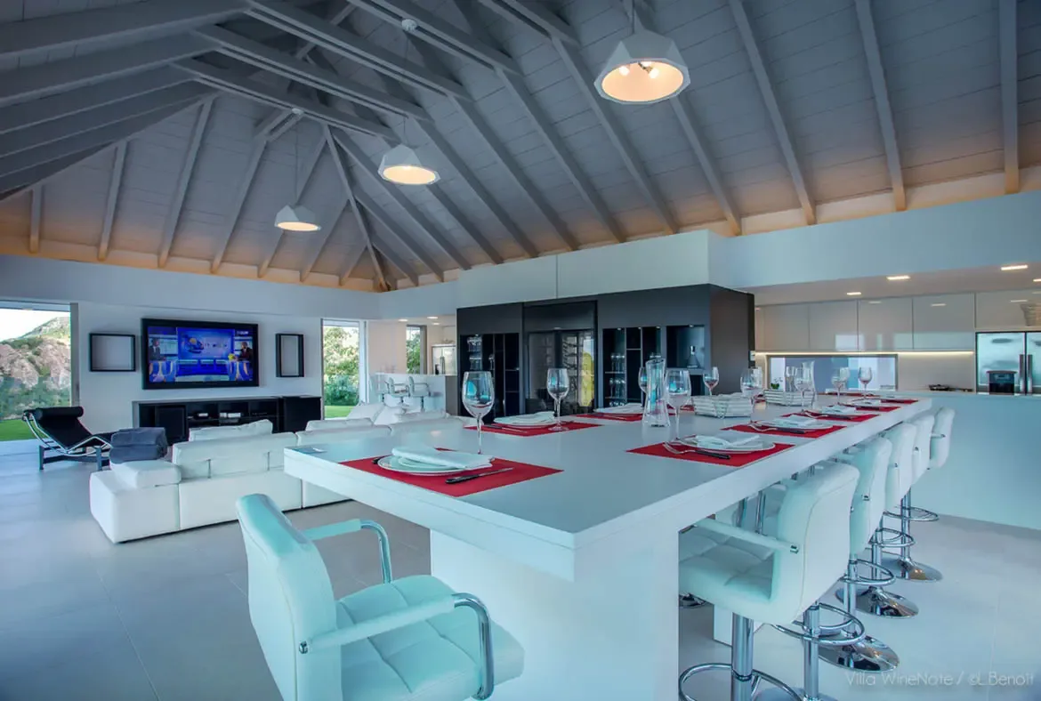 A white dining island set with red placemats and wine glasses sits beside a dark wine display cabinet, where white pendant lights hang from vaulted grey-beam ceilings above a minimalist living space.