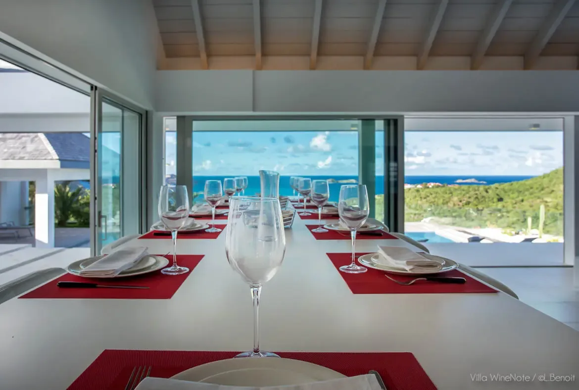 A white dining table set with red placemats and wine glasses sits before open glass walls, where a deep blue ocean panorama fills the vaulted-ceiling dining room with natural light.