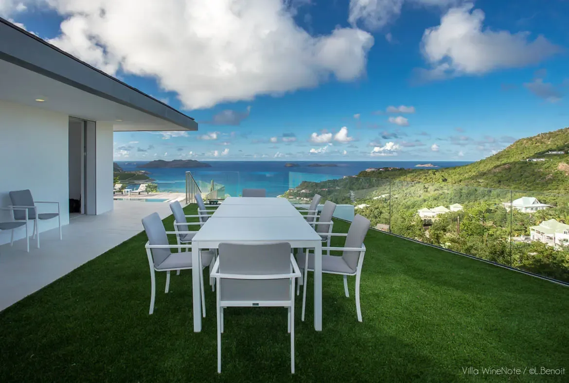 A white dining table with grey chairs sits on green turf beside a modern white villa, where a glass railing opens to a panoramic view of green mountains, a turquoise bay, and distant islands.