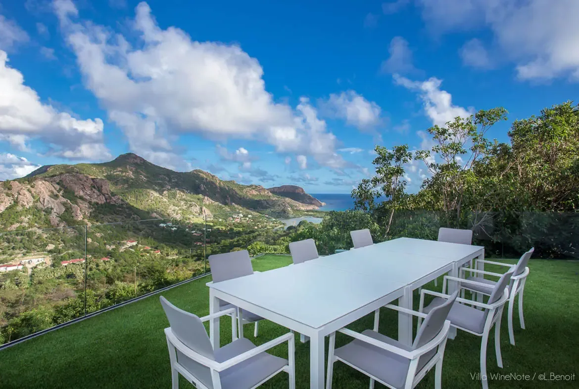 A white dining table with grey-cushioned chairs sits on artificial green turf beside a glass railing, where a lush valley and turquoise bay with islands stretch beneath puffy white clouds.