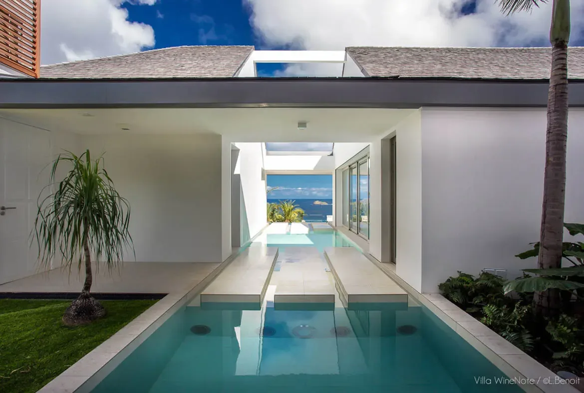 A shallow reflection pool with stone stepping stones leads through a white breezeway framed by palm trees, where the deep blue ocean and distant islands stretch beneath a bright sky.