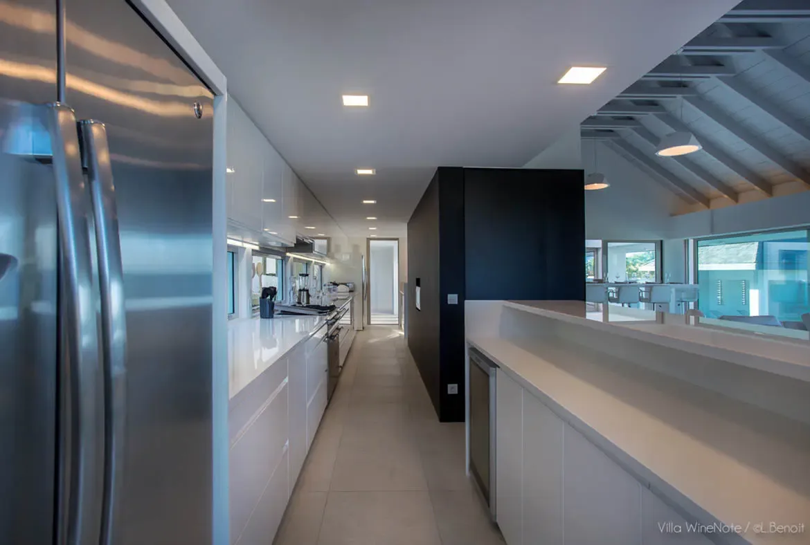 A sleek white kitchen with stainless steel fridge and integrated appliances stretches beside a matte black divider wall, where a wine cooler and recessed lighting accent the minimalist galley.