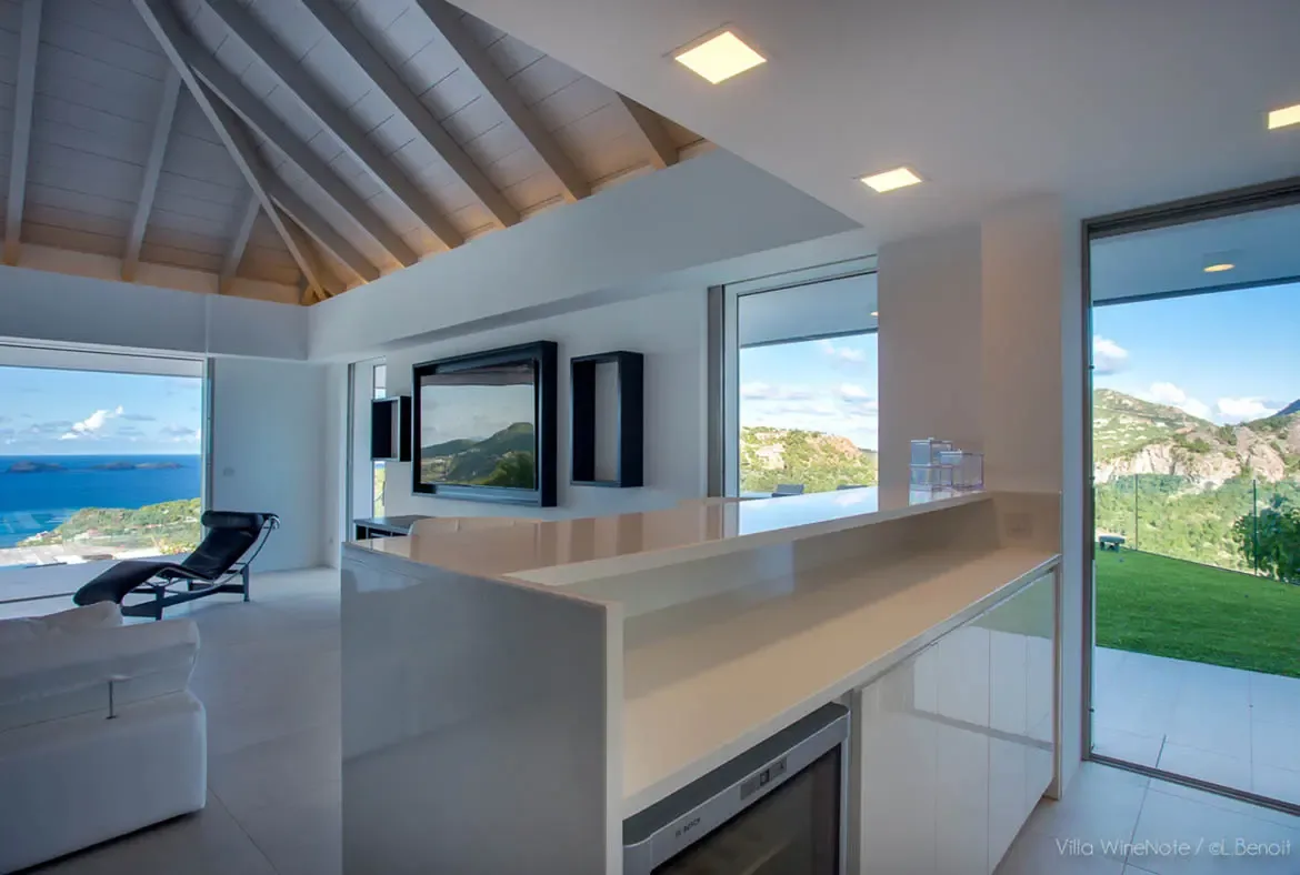 A white bar island with a wine cooler sits beside a black leather chaise and a wall-mounted TV, where open glass doors frame a panoramic ocean view with distant islands.