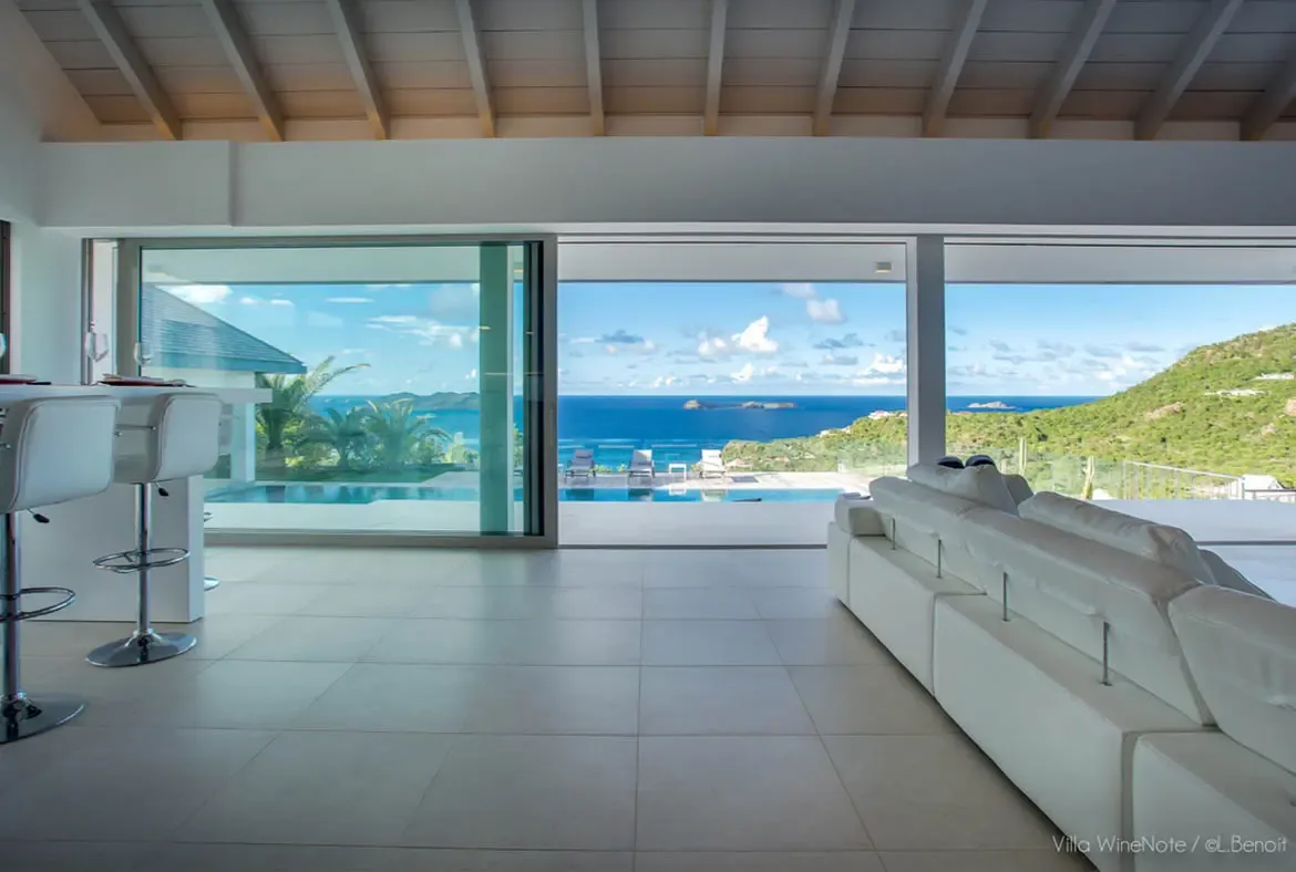 A white sectional sofa faces open glass walls beside a breakfast bar with white stools, where an infinity pool meets a turquoise ocean beneath rolling green hills.