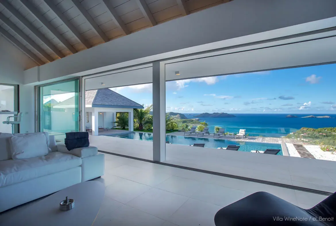 A white sectional sofa sits before floor-to-ceiling glass in a vaulted-beam living room, where an infinity pool and a deep blue ocean with distant islands stretch beneath puffy clouds.