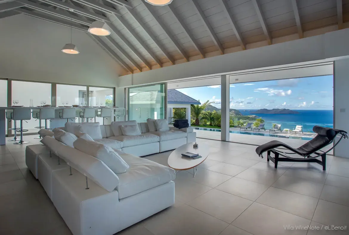 A white sectional sofa and a black leather chaise lounge sit before open glass walls, where an infinity pool and turquoise Caribbean sea with islands fill the panoramic view.
