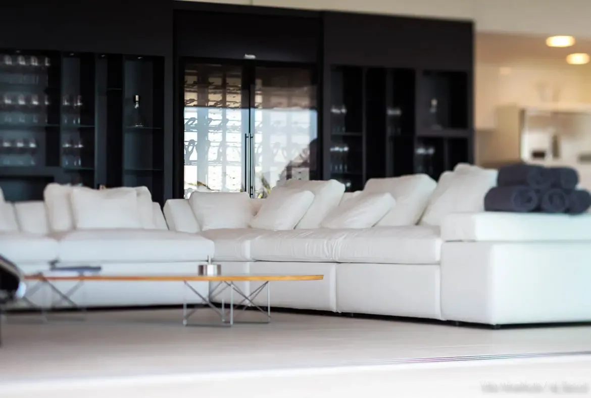A white modular sofa sits before a matte black wall with a walk-in wine cellar and glass display shelves, where warm pendant lights glow above a minimalist living space.