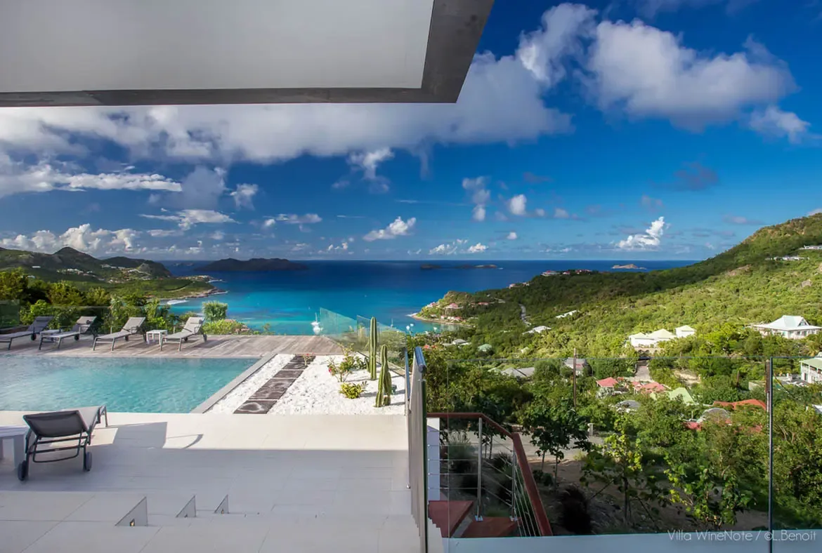 An infinity pool with turquoise water sits beside white loungers on a tiled terrace, where a cactus garden and glass railing overlook a turquoise bay with anchored boats beneath puffy cumulus clouds.