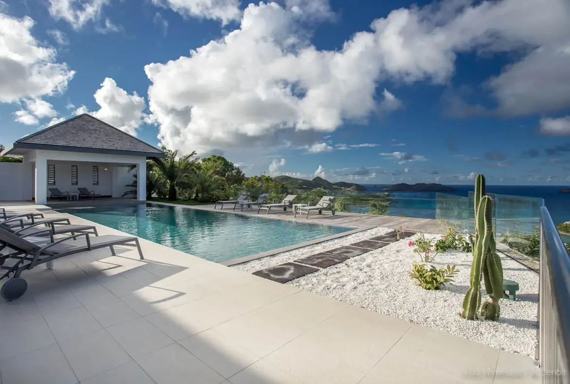 Grey loungers beside an infinity pool sit near a cactus garden and a white cabana, where a frameless glass railing opens to a deep blue ocean with distant islands beneath puffy clouds.