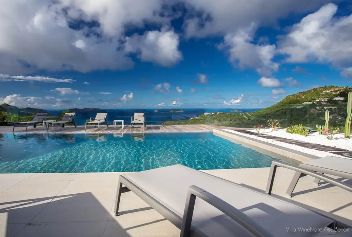Grey loungers beside an infinity pool sit on a light-tiled terrace with a cactus garden, where a deep blue sea dotted with islands stretches beneath dramatic cumulus clouds.