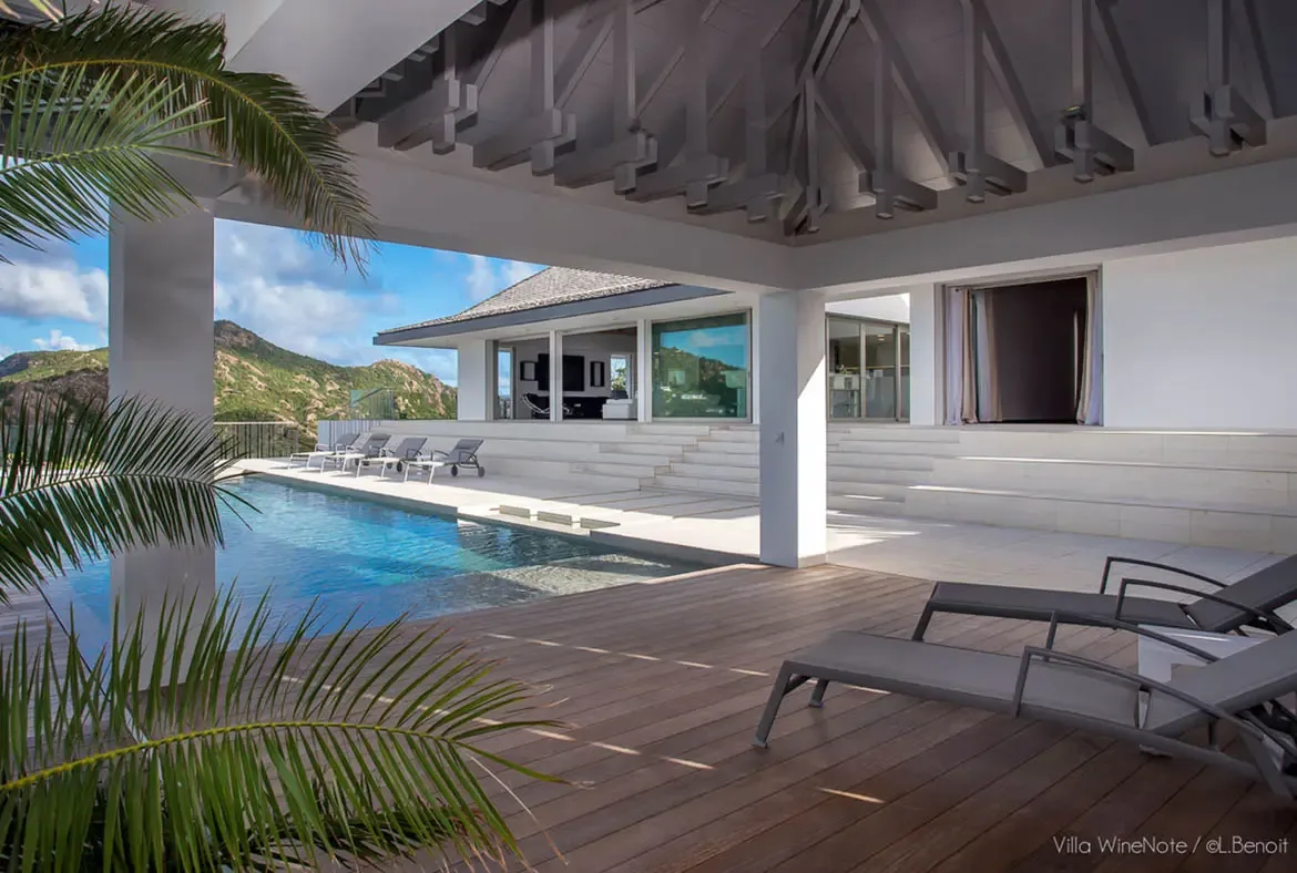 Grey loungers on a wood deck overlook an infinity pool with stone steps, where vaulted wood beams and white pillars frame a panoramic view of lush green mountains beneath a bright sky.