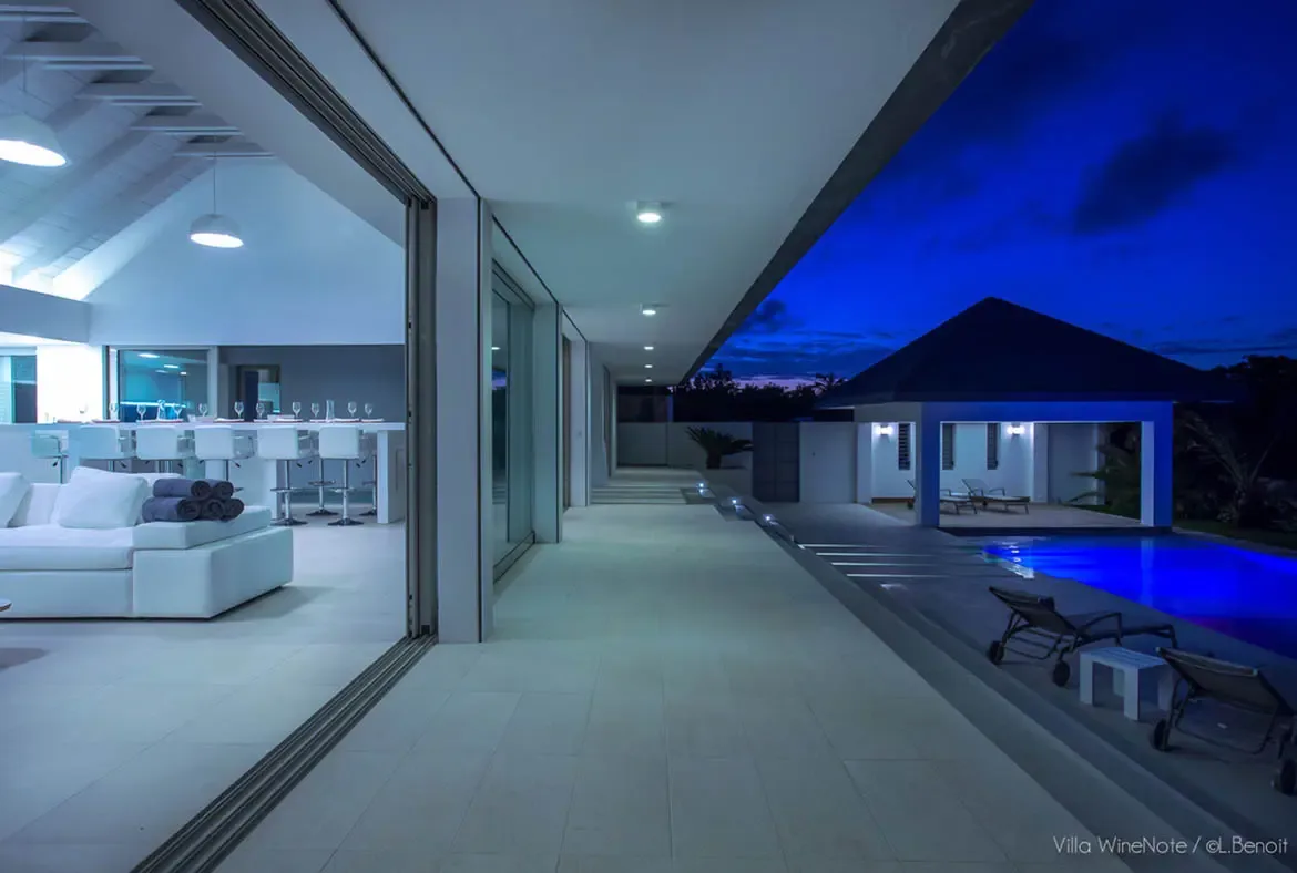A blue-lit infinity pool reflects the villa interior at night, where a white sectional sofa and a dining bar lined with wine glasses glow beneath vaulted white-beam ceilings.