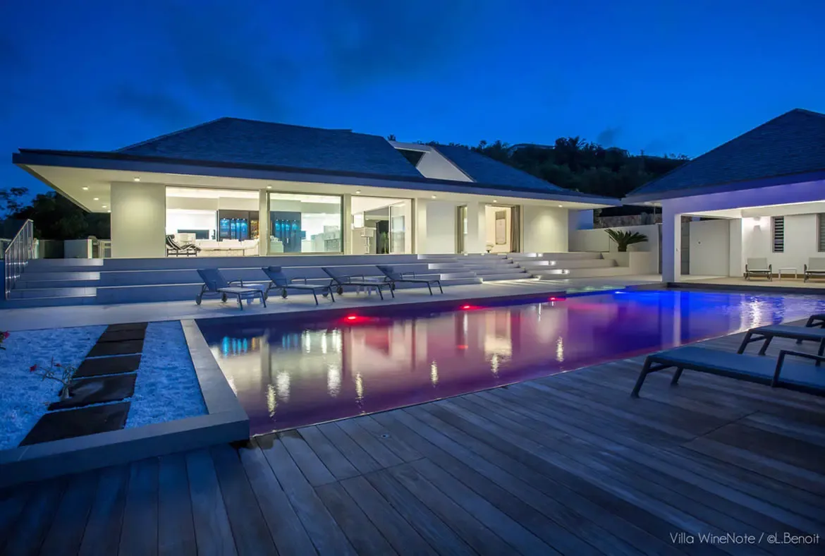 A pool glows with vibrant magenta and blue underwater lights at dusk, where the villa interior reveals an illuminated wine storage area and wide stone steps lead down to the water.