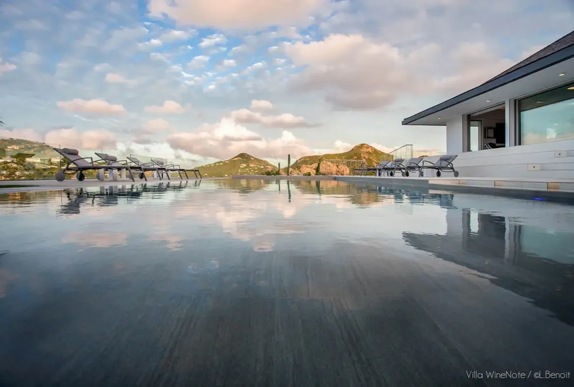 An infinity pool mirrors golden-hour clouds at sunset, where grey loungers and cacti sit on a tiled terrace beneath a hillside dotted with villas.