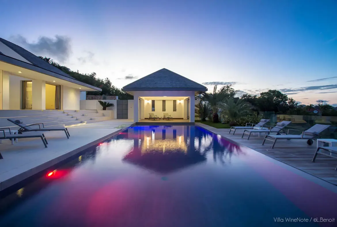 An infinity pool glows with magenta and blue underwater lights at twilight, where a warm-lit pool house pavilion reflects on the water and tiered white steps lead up to the villa.