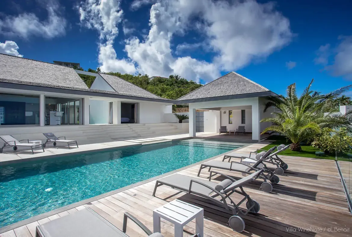 An infinity pool with turquoise water sits beside a wood deck and grey loungers, where a white villa with open glass doors and a palm tree overlooks lush green hills.