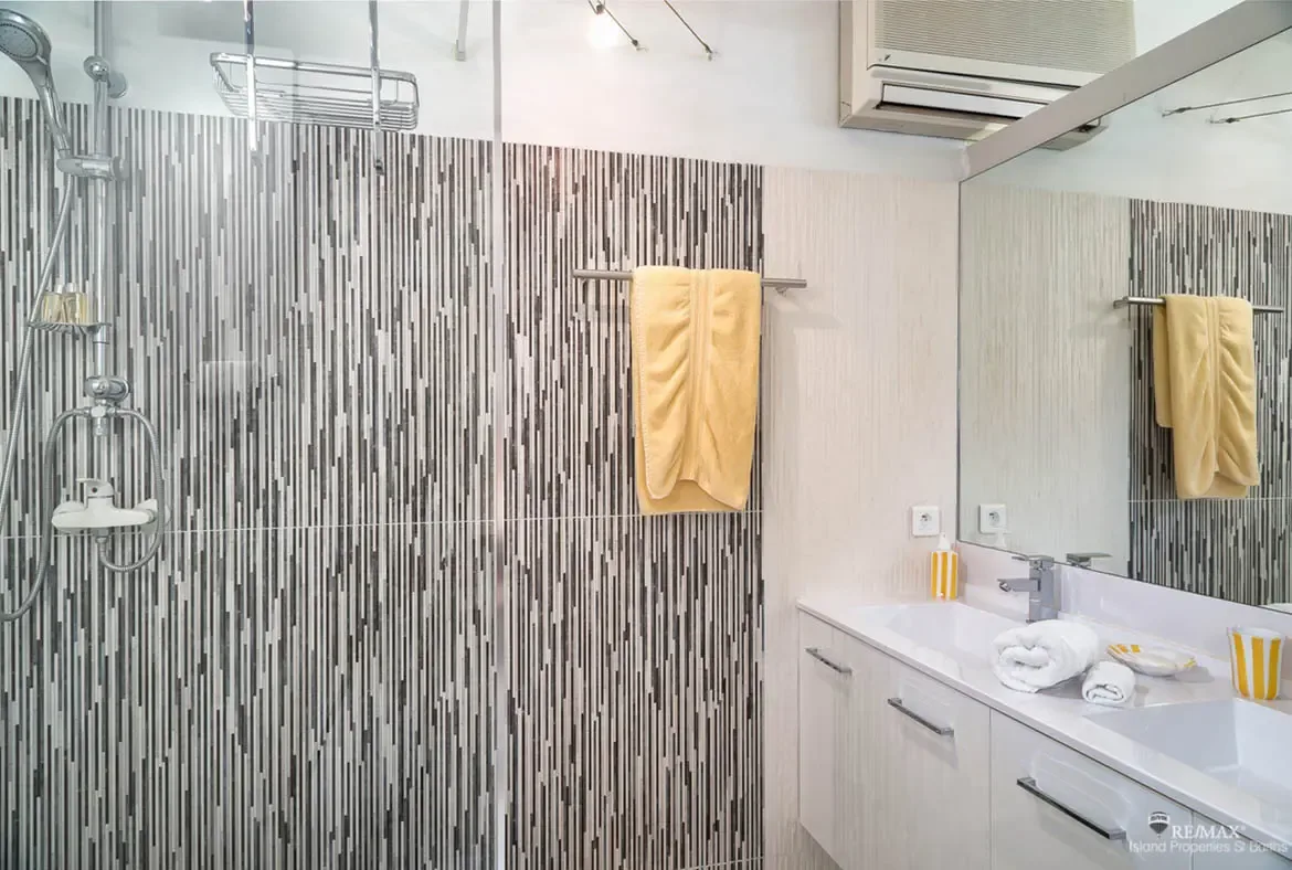 A sleek glass shower with striped gray mosaic tile adds a modern edge to this bright bathroom, while bold yellow towels bring a cheerful splash of color. Clean lines, glossy finishes, and fresh white surfaces keep the space polished and inviting.