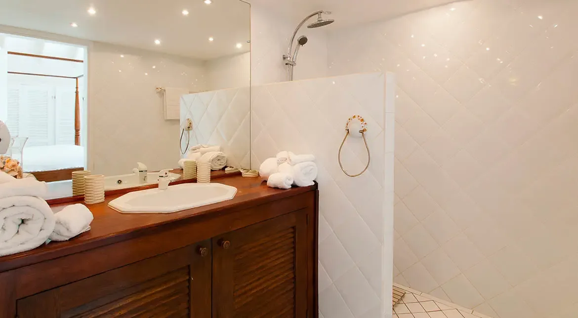 A warm wood vanity with louvered doors and a white oval sink brings texture to this bright bathroom wrapped in glossy white tile. Rolled towels and soft neutral accessories keep the look polished and inviting, with a quiet boutique-hotel feel.