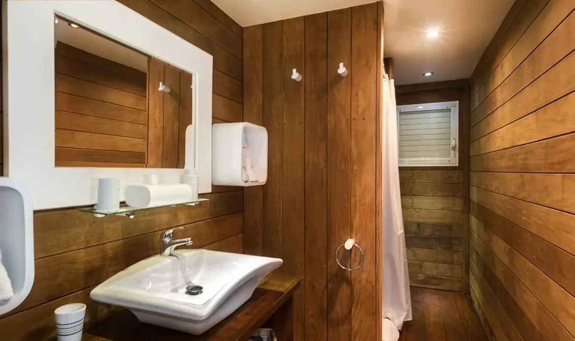 Warm horizontal wood planks wrap this cozy bathroom from floor to ceiling, framing a white vessel sink and crisp white-framed mirror. Built-in niches and a soft shower curtain keep the space serene and uncluttered—a spa-like retreat with tropical warmth.
