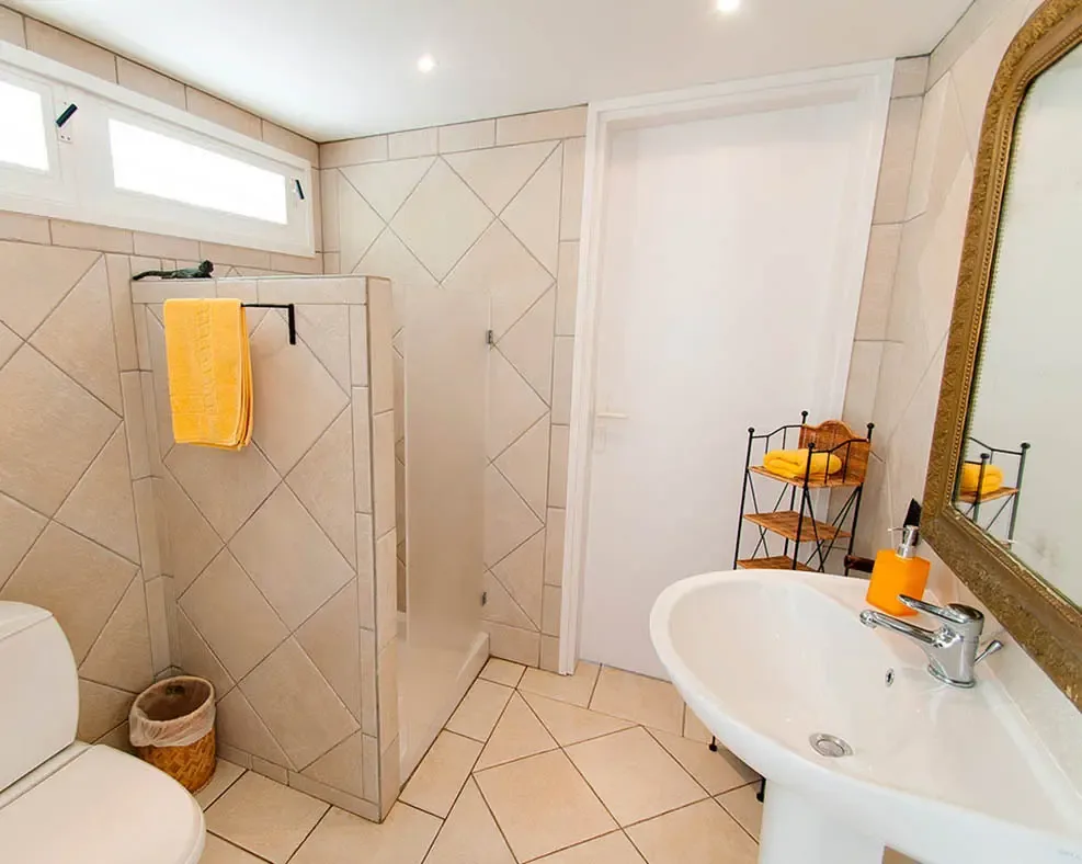 A bright beige-tiled bathroom pairs a glass shower and white pedestal sink with cheerful yellow towels for a fresh tropical touch. An ornate gold-framed mirror and woven storage accents bring warmth and character to the space.