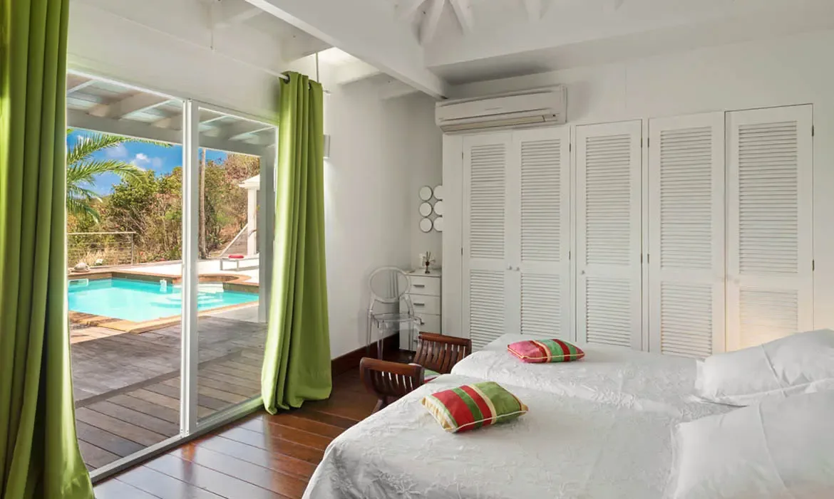 Crisp white linens and striped accent pillows set a cheerful tone in this airy bedroom with exposed white beams and dark wood floors. Green curtains frame the doors to a private deck and turquoise pool—wake up and step straight into vacation mode.