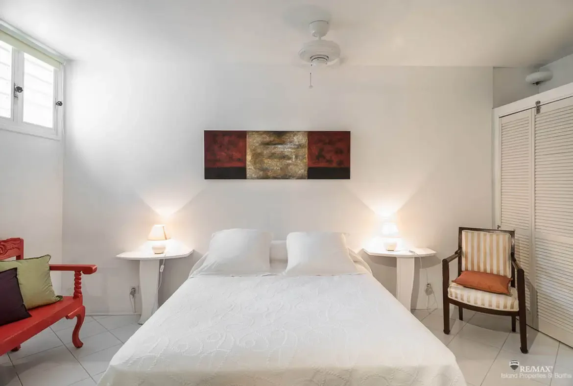 A crisp white bed sits beneath a bold dark artwork, flanked by matching lamps and simple nightstands for a calm, balanced feel. A red bench with colorful pillows brings a cheerful island accent to the room, adding warmth and personality.