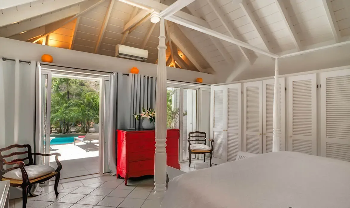 A white four-poster bed sits beneath vaulted beams and a ceiling fan in this airy bedroom, while a bold red dresser adds a playful island accent. Just beyond the doors, the pool and tropical greenery create a peaceful backdrop for slow mornings and restful nights.