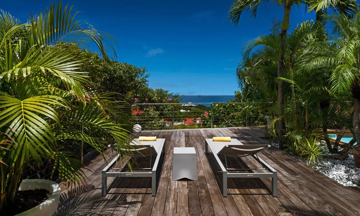 Two sleek loungers with sunny yellow towels sit on a dark wooden deck above the Caribbean, framed by palms and a glimpse of the pool below. Red rooftops peek through the greenery while the ocean stretches endlessly beyond—an easy spot to spend the entire afternoon.
