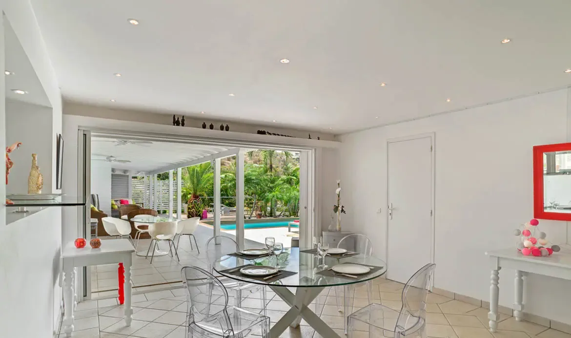 A round glass dining table set for four sits between bright white walls, clear acrylic chairs, and colorful decorative accents. Beyond the sliding doors, the covered patio opens to the pool and tropical greenery, making every meal feel connected to the outdoors.