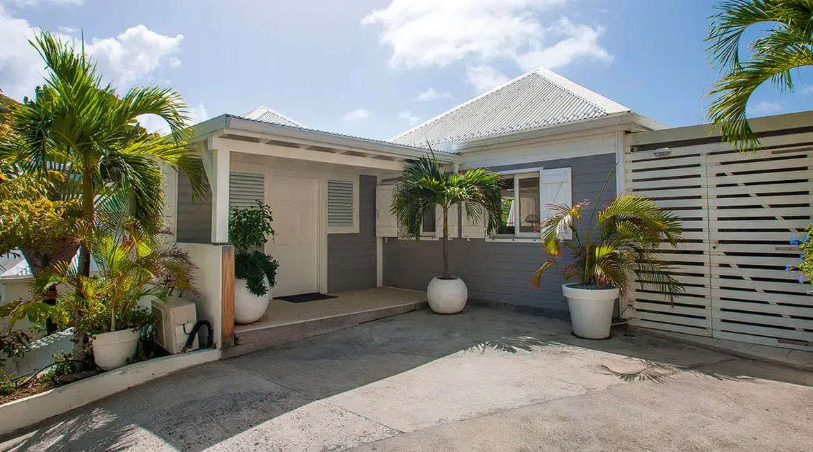 A light gray villa with crisp white trim and a metal roof sits behind a clean driveway framed by oversized white planters and swaying palms. Tropical greenery softens every edge, giving the entrance an easy, elegant island feel.
