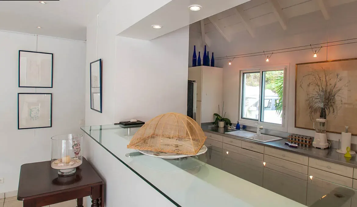 A glass-topped bar anchors this bright open kitchen with white cabinetry, a vaulted beamed ceiling, and clean contemporary lines. Beach-inspired decor, soft lighting, and curated details give the space an easy elegance for slow breakfasts and evening cocktails alike.