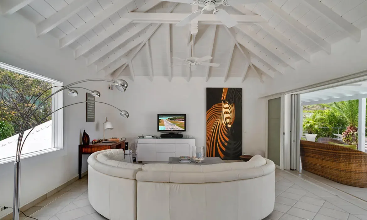 A curved white leather sectional sits beneath vaulted white beams and twin ceiling fans in this bright, open living room. Bold zebra artwork adds personality while the open doors and greenery outside keep the whole space feeling breezy and relaxed.