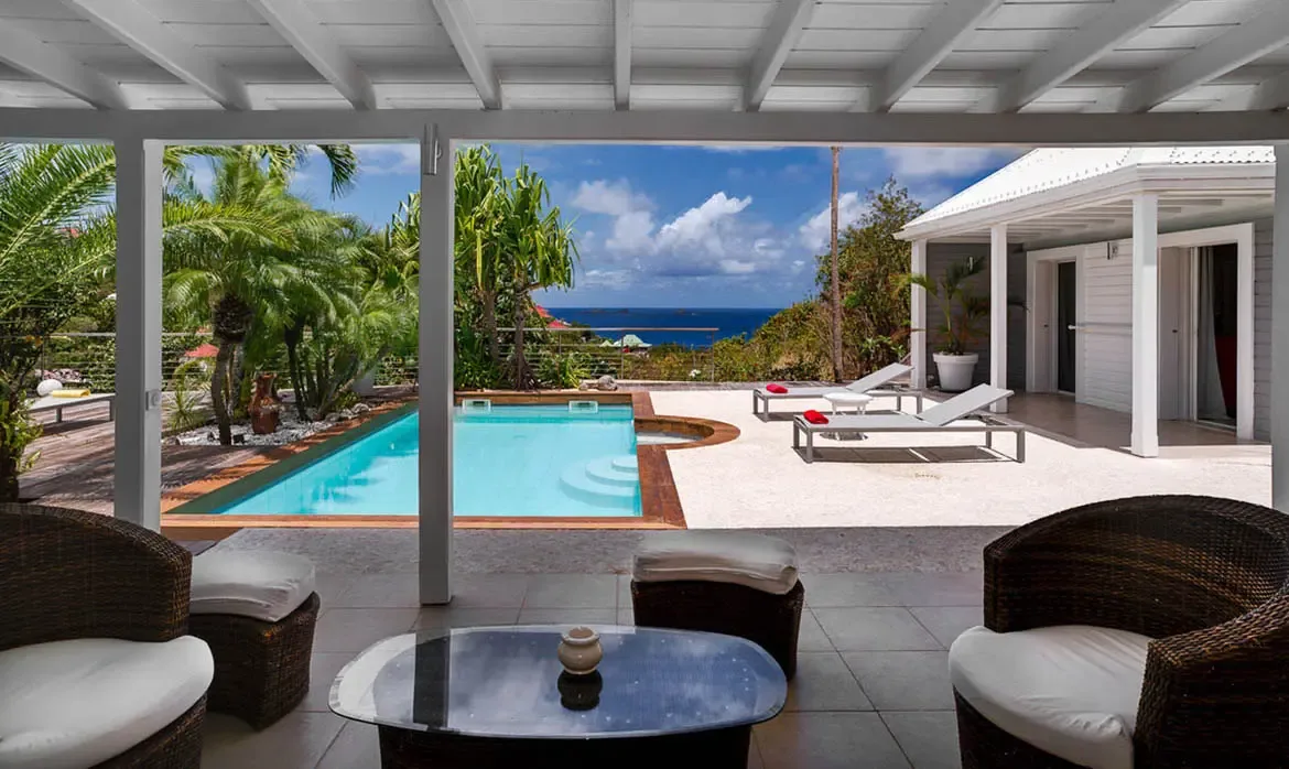 A shaded patio with wicker armchairs and a glass-topped table opens onto a curved turquoise pool and a stone deck with red-cushioned loungers. Palms, ocean blue, and white columns complete the scene, creating an easy flow from morning coffee to poolside afternoons.