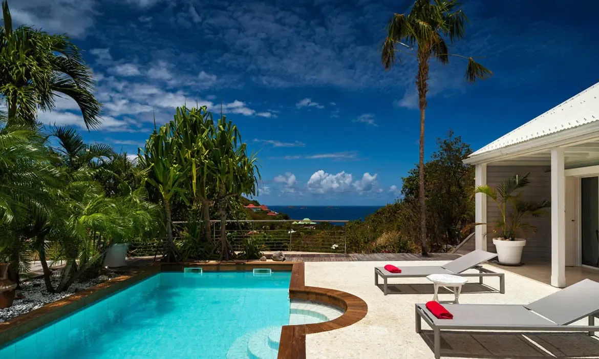 A turquoise pool with a curved inset lounge area stretches toward the ocean while two gray loungers with vivid red towels invite sun-soaked afternoons. Framed by palms and a white-roofed villa, this is classic St. Barths poolside bliss.