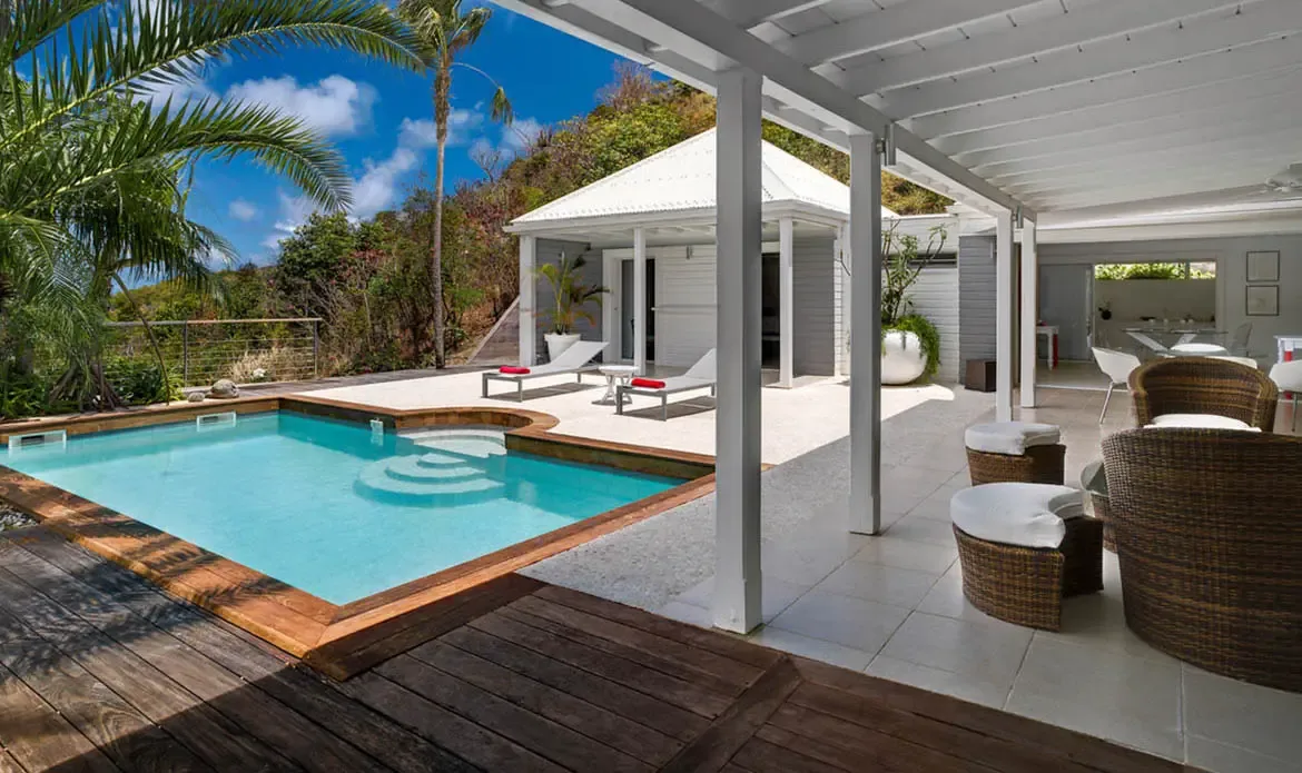 A turquoise pool curves alongside a dark wooden deck and a shaded patio filled with wicker seating and white cushions. The white villa opens wide to the terrace, surrounded by palms and tropical hills for an effortless indoor-outdoor island retreat.