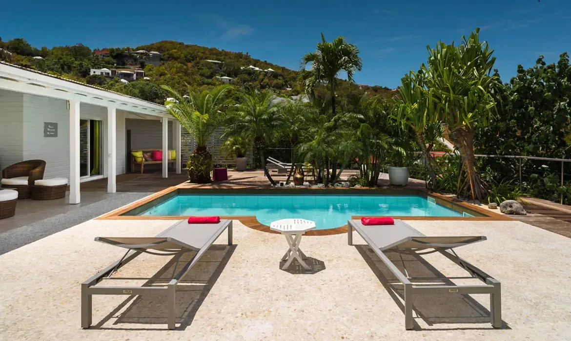 A crystal-clear pool stretches across the terrace where two loungers with bright red towels face the sun. Beneath the shaded patio, wicker seating with colorful pillows adds a playful island touch while palms and hillside greenery surround the scene.