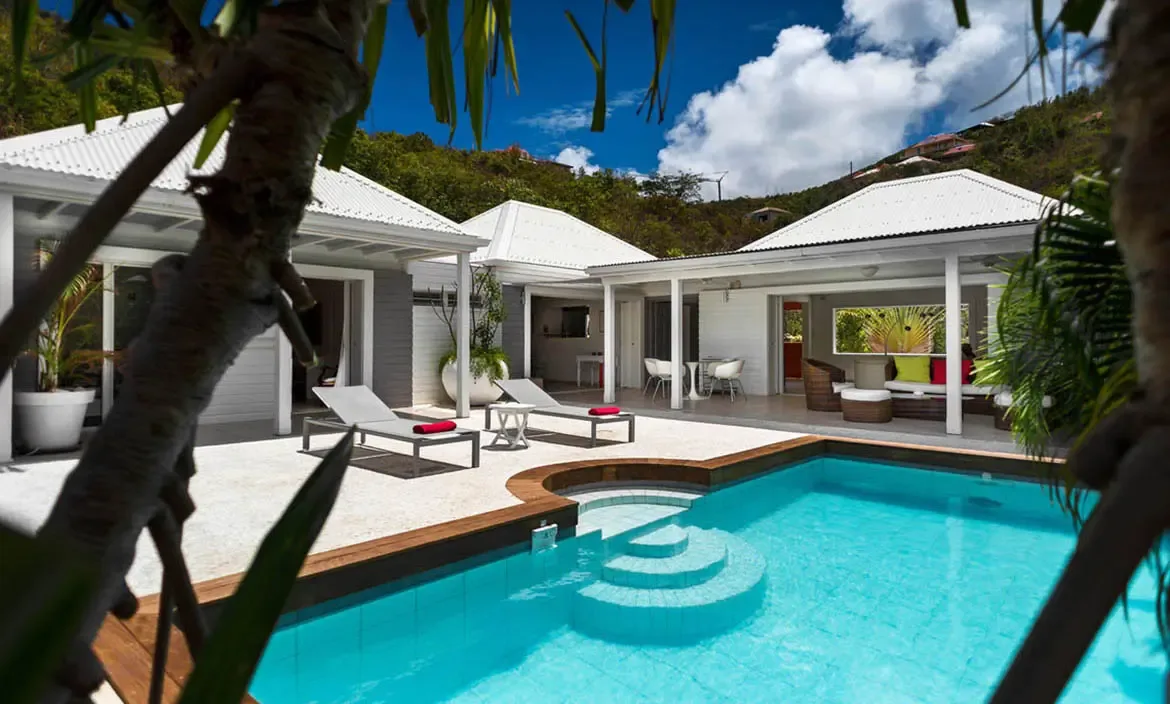 A turquoise pool with a rounded inset section anchors this breezy villa compound of white-roofed pavilions and shaded terraces. Red-cushioned loungers and colorful outdoor seating bring playful energy to the scene, all set against the lush St. Barths hillside.