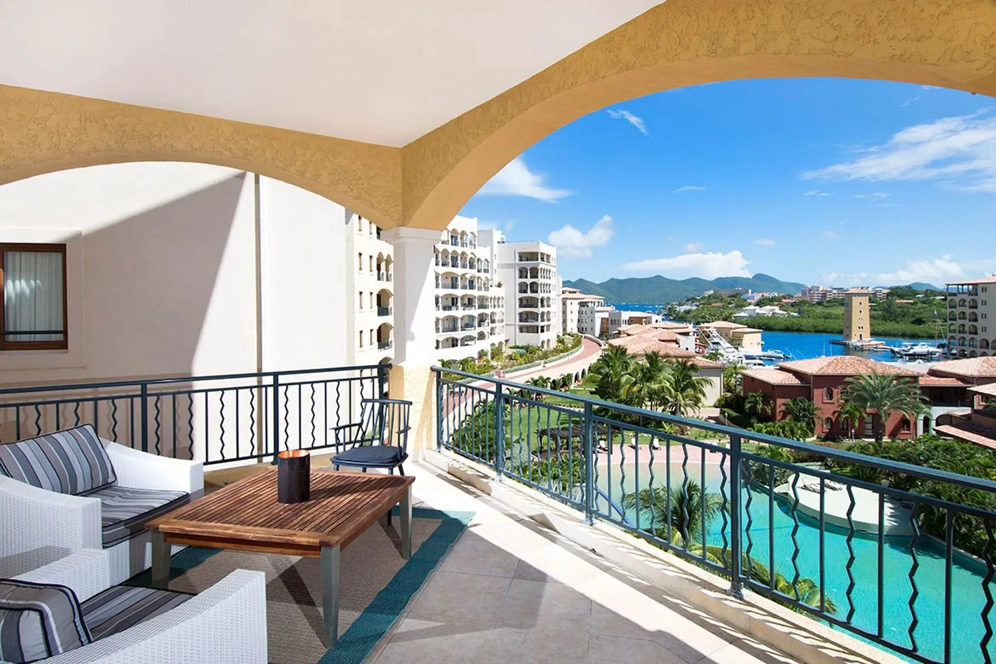 A sun-drenched balcony with elegant arched openings reveals sweeping marina vistas — white yachts bobbing on sapphire waters below.