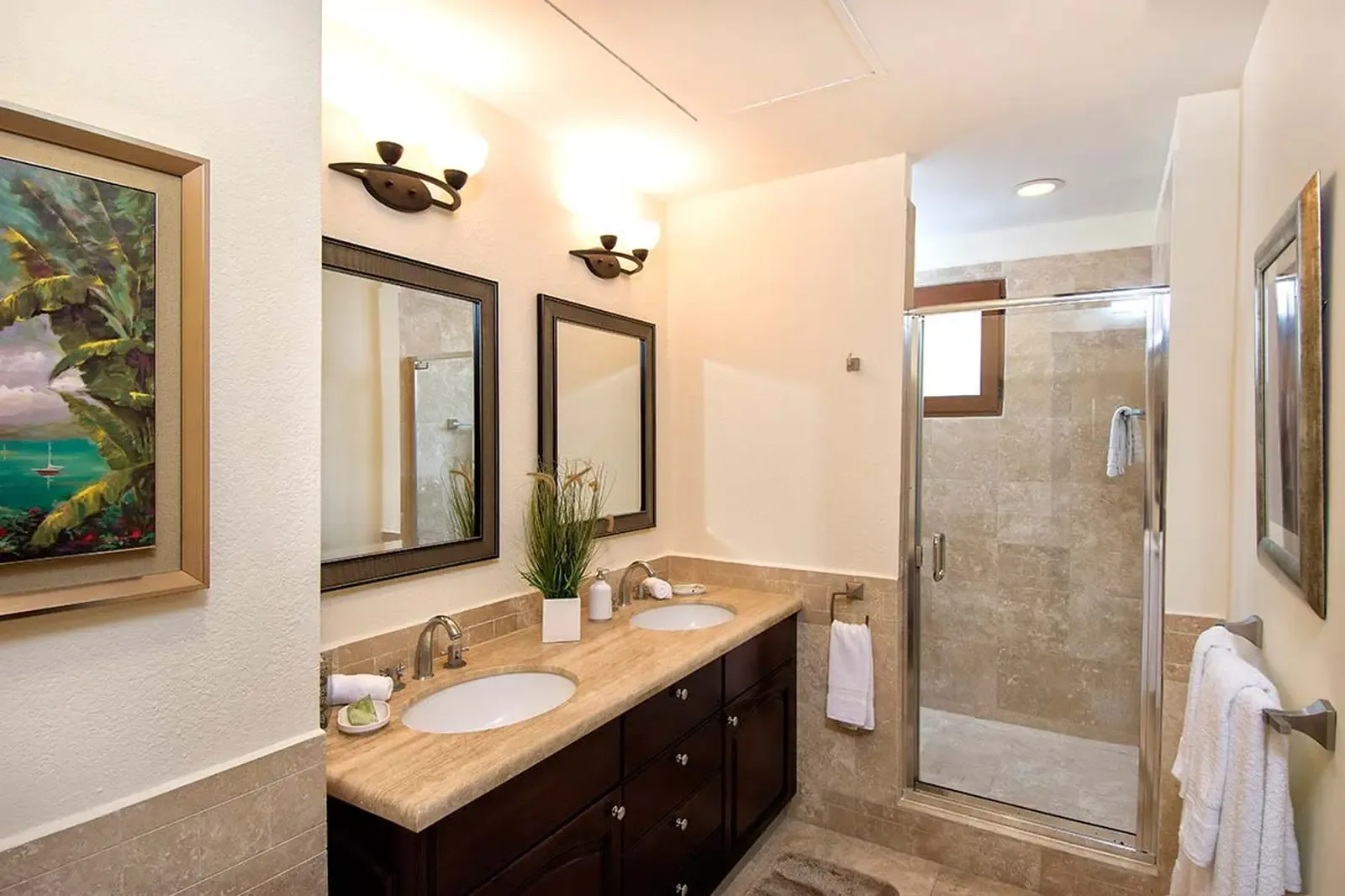 A travertine-clad double vanity and espresso cabinetry define a spa-like ensuite, softened by warm brass sconces and lush tropical artwork.