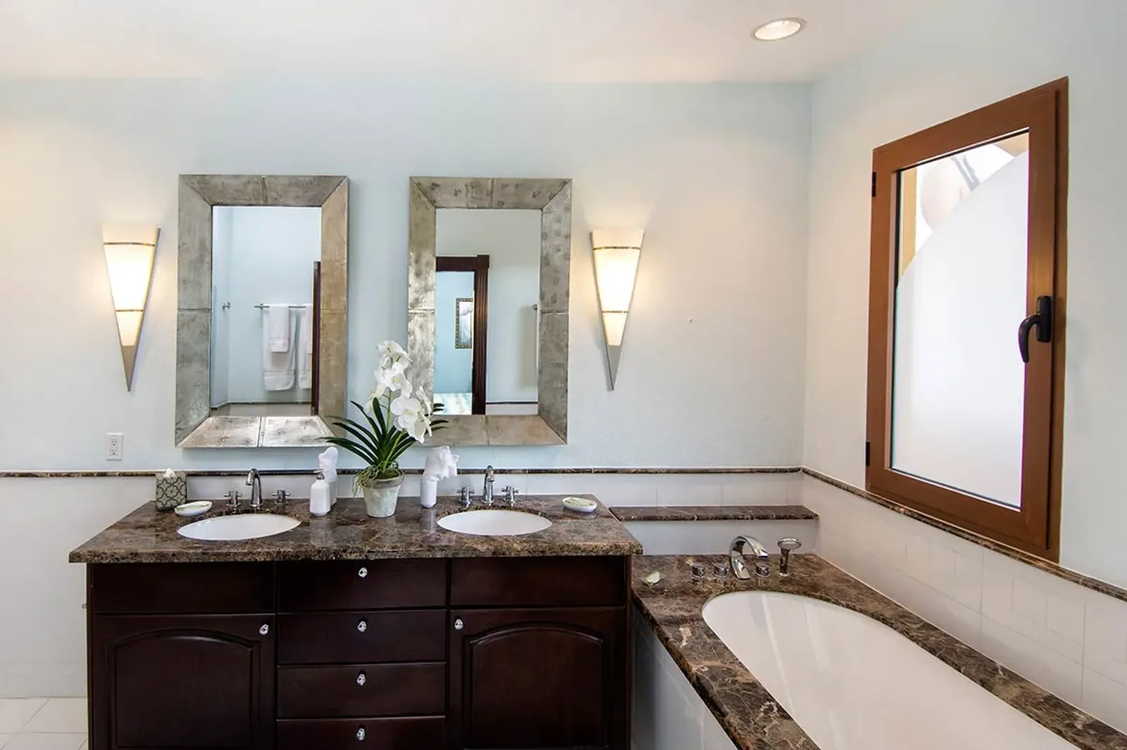 Emperador marble wraps a deep soaking tub beneath a frosted casement window — a private spa where warm stone meets cool chrome fixtures.