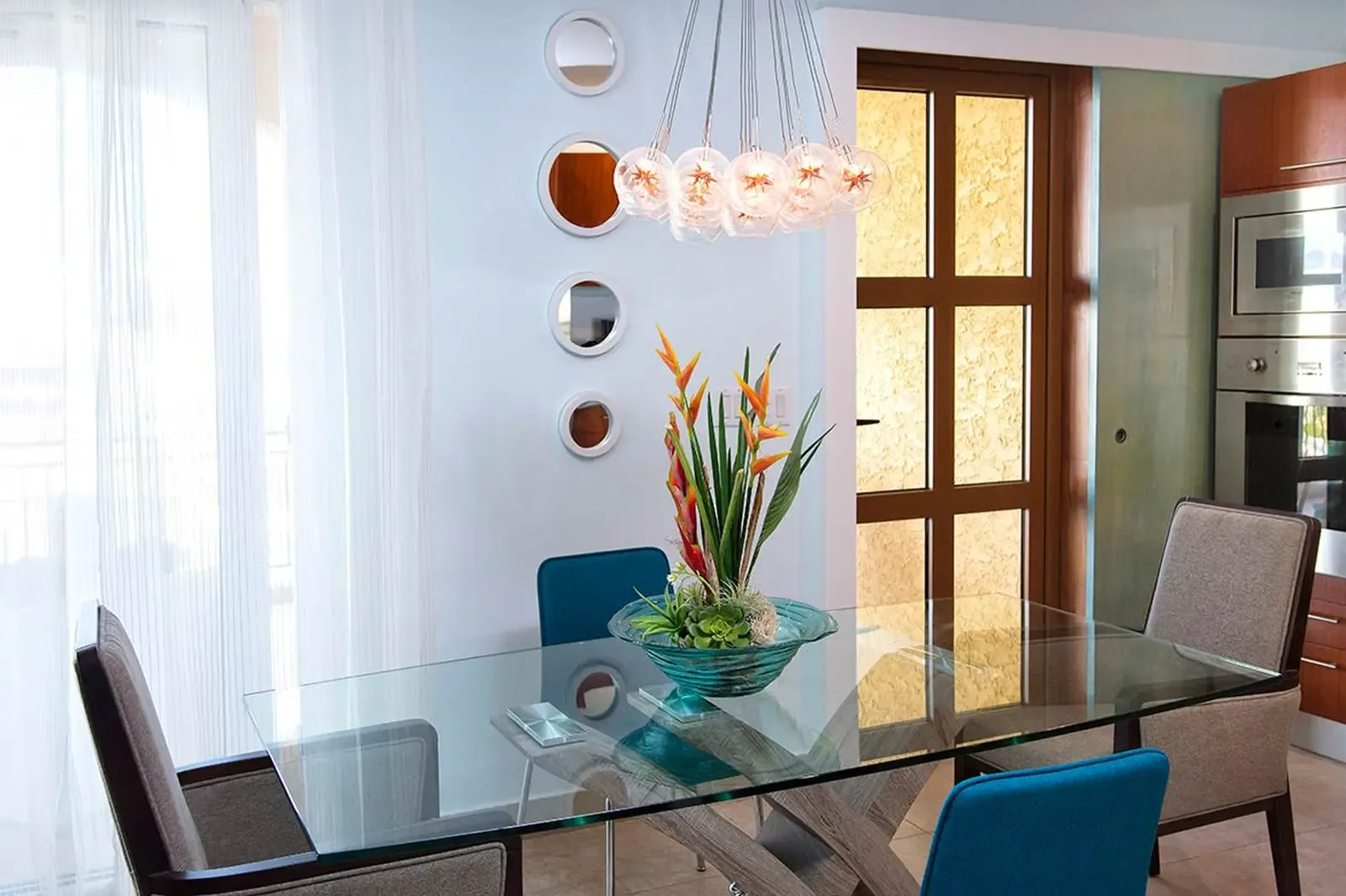 A glass-top dining table reflects the glow of a multi-sphere chandelier, surrounded by teal-upholstered chairs and Birds of Paradise in turquoise glass.